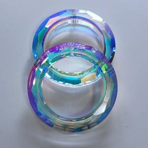May include: Two circular, faceted crystal rings featuring an iridescent finish that reflects shades of blue, purple, and green. Each ring has a smooth, clear center opening and a multi-faceted outer edge that catches light to create a prismatic effect.