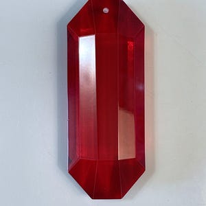 May include: A faceted, ruby-red crystal pendant with a rectangular shape and a small hole at the top. The pendant has a geometric design with multiple flat surfaces that reflect light. The crystal is a deep, transparent red.