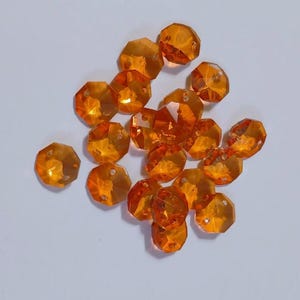 May include: A collection of faceted, orange-colored beads with a hole drilled through each. The beads have a geometric, crystal-like appearance and are scattered on a white surface. These beads are suitable for jewelry making and craft projects.