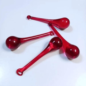 May include: Four red glass teardrop ornaments with a bulbous end and a loop at the top. The ornaments are arranged diagonally against a white background. The glass has a glossy finish.