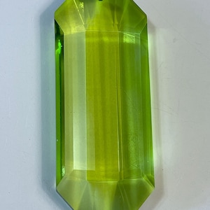 May include: A faceted, elongated, lime green crystal pendant with a hole at the top for hanging. The crystal has a rectangular shape with angled edges and a polished, transparent appearance. The pendant is approximately 3 inches long.