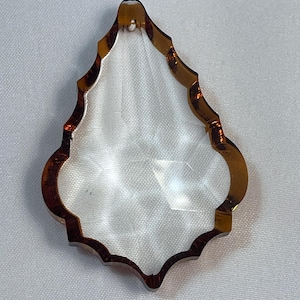May include: A clear, teardrop-shaped crystal pendant featuring a faceted surface and a distinct amber-colored border. The piece has a pointed top with a small hole for suspension and a scalloped, decorative edge that frames the transparent center.