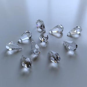 May include: A collection of ten clear, faceted crystal beads shaped like teardrops. Each bead features multiple geometric surfaces that reflect light, creating a sparkling appearance. The beads are arranged in a scattered pattern on a smooth surface.