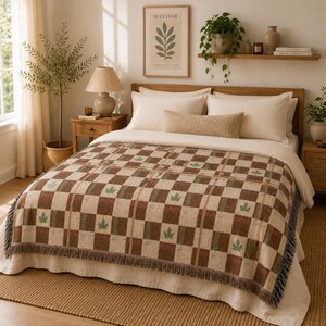Könnte beinhalten: Eine gewebte Wohndecke mit einem braun-cremefarbenen Karomuster und kleinen grünen Blattmotiven. Die Decke hat eine strukturierte Oberfläche und Fransen an den Rändern und liegt auf einem Bett mit weißen Kissen und einem beigen Kissen.