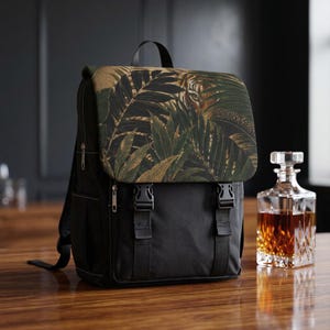 May include: A black backpack featuring a top flap with a dark green and gold tropical leaf pattern and a hidden tiger face. The bag includes two black plastic buckle closures on the front, a top handle, and side pockets with silver-toned zippers.