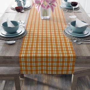 May include: A rectangular table runner featuring a plaid pattern in shades of orange, white, and dark green. The runner is centered on a wooden table, with place settings including blue bowls, white napkins, and wine glasses on each side.