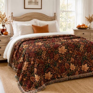 May include: A woven throw blanket featuring a dark, multi-coloured pattern of autumn leaves, acorns, and berries in shades of deep red, brown, green, and gold. The blanket has a fringed edge and is draped over a bed with white and orange pillows.
