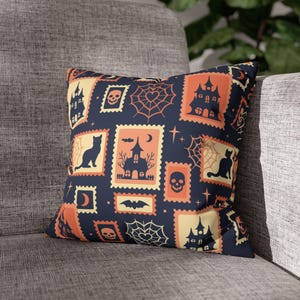 May include: A square throw pillow featuring a dark blue background with a repeating pattern of rectangular postage stamp-style illustrations in orange and cream. The designs include black silhouettes of haunted houses, cats, skulls, bats, and spider webs.