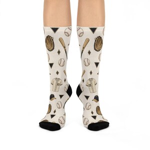 May include: A pair of off-white crew socks featuring a repeating pattern of baseball equipment including brown gloves, wooden bats, white baseballs, baseball jerseys with numbers, and black caps, interspersed with small black triangles and diamonds.