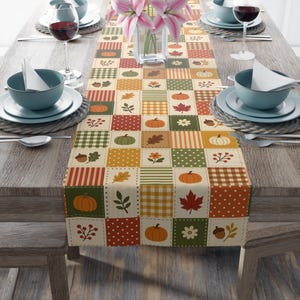 May include: A rectangular table runner featuring a patchwork design of autumn-themed squares. The pattern includes illustrations of orange pumpkins, red maple leaves, brown acorns, green foliage, and white flowers, interspersed with plaid and polka dot squares.