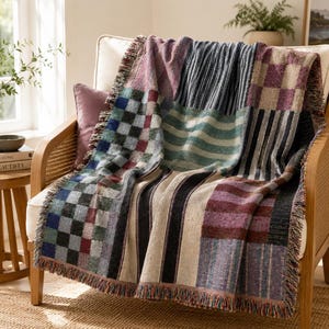 Puede incluir: Una manta tejida con un diseño de retazos que presenta varios patrones, incluyendo cuadrados a cuadros, rayas verticales y bandas horizontales en tonos apagados de azul, morado, crema y verde oscuro con un borde de flecos en la parte inferior.