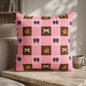 May include: A square throw pillow featuring a checkered pattern of alternating light pink gingham squares and solid dark brown squares. The dark brown squares are decorated with repeating motifs of dark brown bows, light tan bows, and brown maple leaves.