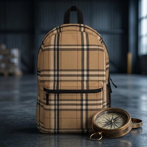 May include: A tan backpack featuring a black, white, and red plaid pattern with a front zippered pocket and a top handle. Positioned in front of the backpack is a round, antique-style brass compass with a detailed face and a metal ring attachment.