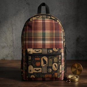 May include: A backpack featuring a brown, green, and cream plaid pattern on the main body. The front pocket displays a dark print with motifs including a black cat, a pumpkin, bats, spiders, moons, and birds. A brass pocket watch sits nearby.