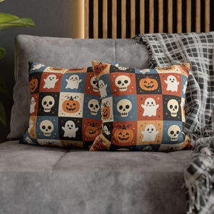 以下が含まれることがあります： ハロウィーンをテーマにしたイラストが格子状に繰り返される、2つの正方形のクッション。赤、青、黒の四角い枠の中に、白いお化け、オレンジ色のジャック・オー・ランタン、ベージュ色のドクロが描かれたデザインが特徴です。