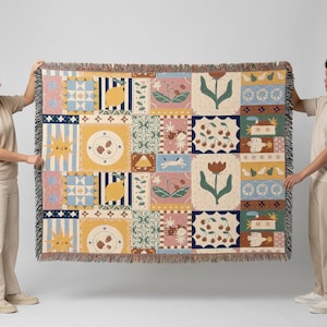 May include: A rectangular woven throw blanket featuring a multi-panel grid design with various illustrations including lemons, cherries, flowers, suns, and small houses in a palette of yellow, blue, pink, green, and brown with fringed edges.