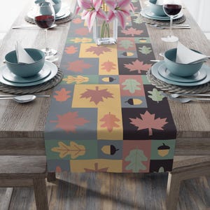 May include: A rectangular table runner featuring a repeating grid pattern of autumn-themed motifs including maple leaves, oak leaves, and acorns in shades of muted yellow, sage green, dusty rose, and charcoal grey, centred on a wooden dining table.