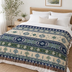 May include: A woven throw blanket featuring a cream-colored base with horizontal bands of navy blue and forest green. The pattern includes geometric shapes, star-like motifs, and stylized tree silhouettes, finished with a fringed edge on the ends.