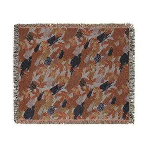 May include: A rectangular woven throw blanket featuring a repeating pattern of dark silhouettes of turkeys, brown maple leaves, and pinecones against a textured rust-orange background. The blanket has a decorative fringed edge on all four sides.