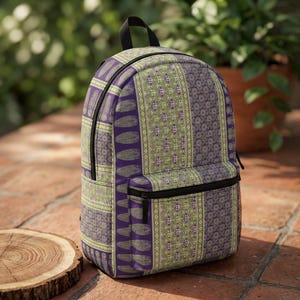 May include: A backpack featuring a multi-patterned design in shades of purple and light green. The fabric displays vertical panels with repeating feather motifs, geometric diamond shapes, and intricate floral-like patterns, finished with black zippers.