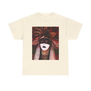 May include: A t-shirt featuring a woman with a red and brown leafy headdress and a black mask over her eyes. Her face is painted white with black accents around her eyes.
