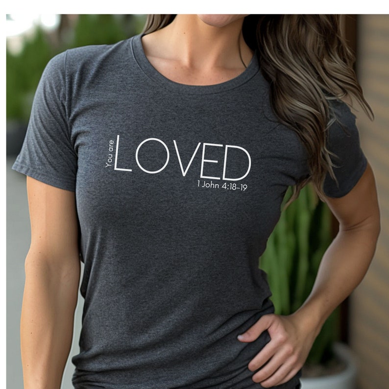 Może przedstawiać: Szary t-shirt z nadrukiem słowa "LOVED" w kolorze białym, a także tekstem "You are" i "1 John 4:18-19".