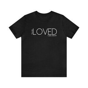 Może przedstawiać: Czarny T-shirt z białym napisem "You are LOVED 1 John 4:18-19".