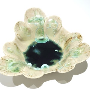 May include: A ceramic bowl with a unique, scalloped edge design. The bowl features a dark blue center, surrounded by cream-colored edges with green and brown accents. The overall shape is reminiscent of a flower.