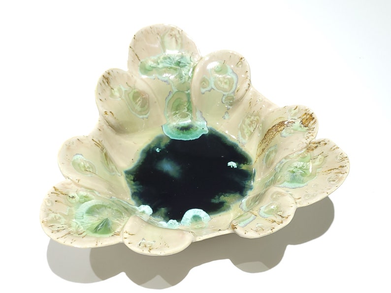 May include: A ceramic bowl with a unique, scalloped edge design. The bowl features a dark blue center, surrounded by cream-colored edges with green and brown accents. The overall shape is reminiscent of a flower.