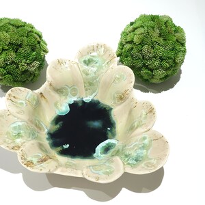 May include: A ceramic bowl with a dark blue center and a cream-colored rim with green accents. Two green, textured spheres are positioned on either side of the bowl, all set against a white background.