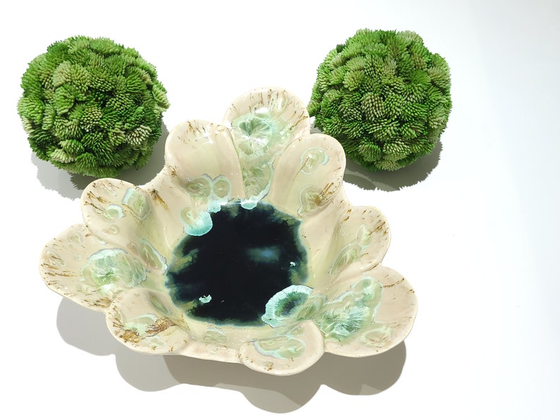May include: A ceramic bowl with a dark blue center and a cream-colored rim with green accents. Two green, textured spheres are positioned on either side of the bowl, all set against a white background.
