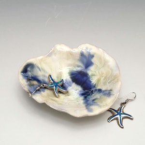 May include: A ceramic seashell-shaped dish with blue and white swirls, holding two starfish earrings. The earrings have silver-colored metal and blue-green centers, with a hook closure. The dish is approximately 6 inches wide.