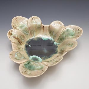 May include: A handmade ceramic bowl with a unique, organic shape. The bowl features a dark blue center and a creamy, off-white exterior with green and brown accents. The edges have a scalloped design, adding to its artistic appeal.