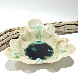 May include: A ceramic bowl with a scalloped edge, featuring a cream-colored exterior with green and gold accents. The interior has a dark blue-green glaze. A piece of driftwood rests behind the bowl.