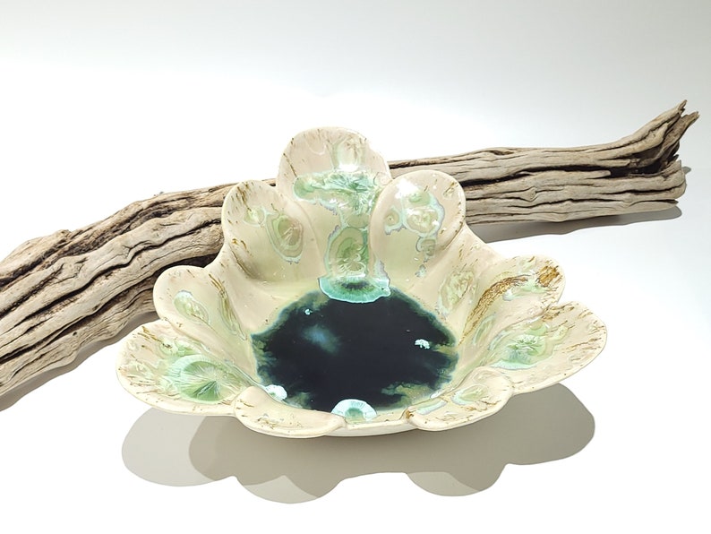 May include: A ceramic bowl with a scalloped edge, featuring a cream-colored exterior with green and gold accents. The interior has a dark blue-green glaze. A piece of driftwood rests behind the bowl.