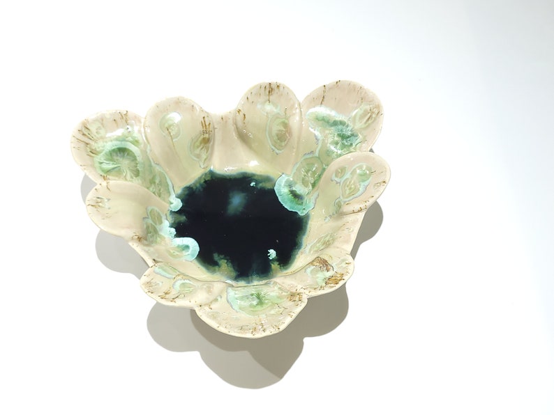 May include: A ceramic bowl with a unique, organic shape. The bowl features a dark blue center and a light beige exterior with green and brown accents. The edges have a scalloped design, adding to its artistic appeal. This decorative bowl is ideal for serving or display.
