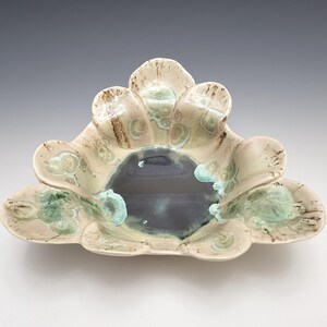 May include: A handcrafted ceramic bowl with a unique, organic shape. The bowl features a central dark blue-gray interior surrounded by petal-like edges in cream, brown, and turquoise hues. The bowl's design is reminiscent of a flower.
