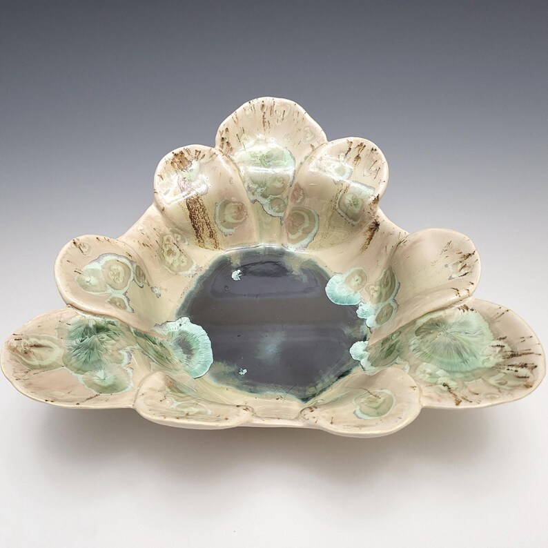 May include: A handcrafted ceramic bowl with a unique, organic shape. The bowl features a central dark blue-gray interior surrounded by petal-like edges in cream, brown, and turquoise hues. The bowl's design is reminiscent of a flower.