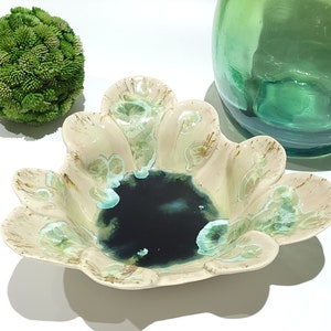 May include: A decorative ceramic bowl with a unique, scalloped edge design. The bowl features a dark blue center and is adorned with light green and brown accents. A green glass vase and a green textured sphere are also in the image.