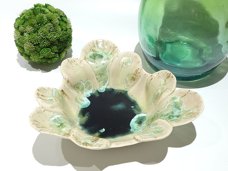 May include: A decorative ceramic bowl with a unique, scalloped edge design. The bowl features a dark blue center and is adorned with light green and brown accents. A green glass vase and a green textured sphere are also in the image.