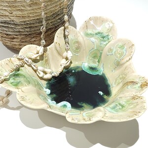 May include: A decorative ceramic bowl with a scalloped edge, featuring a dark blue center and green and cream accents. A seashell necklace and a woven basket are also in the image, creating a coastal theme.