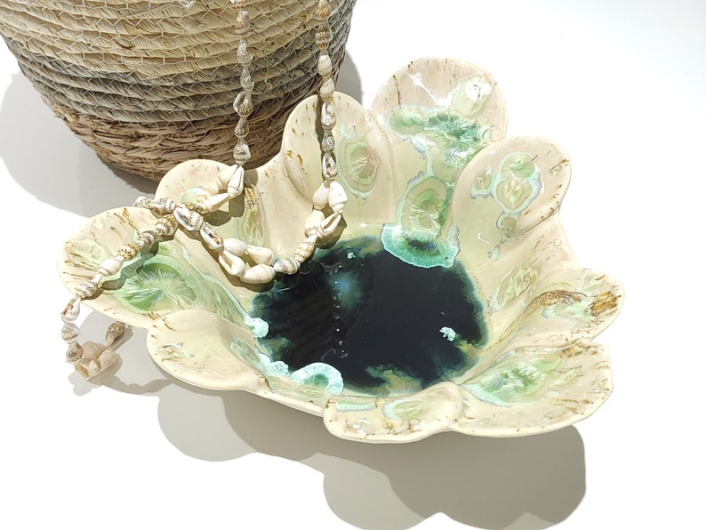 May include: A decorative ceramic bowl with a scalloped edge, featuring a dark blue center and green and cream accents. A seashell necklace and a woven basket are also in the image, creating a coastal theme.