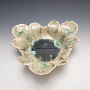 May include: A handcrafted ceramic bowl with a unique, organic shape. The bowl features a dark, glossy interior and a light beige exterior with turquoise and brown accents. The edges are gently scalloped, creating a flower-like appearance. A decorative piece for the home.
