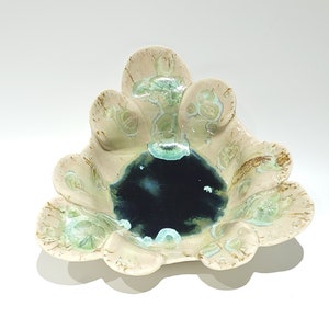 May include: A ceramic bowl with a unique, organic shape. The bowl features a cream-colored exterior with brown and green accents. The center of the bowl is a deep, dark blue. The bowl has a scalloped edge.