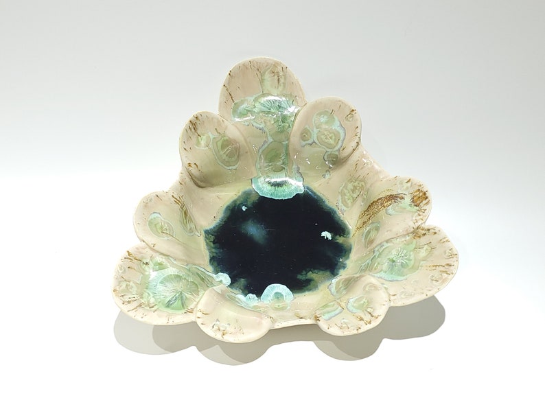 May include: A ceramic bowl with a unique, organic shape. The bowl features a cream-colored exterior with brown and green accents. The center of the bowl is a deep, dark blue. The bowl has a scalloped edge.