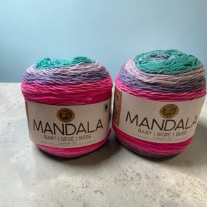 以下が含まれることがあります： 「MANDALA」の文字が印刷された、2つのカラフルな毛糸玉。ピンク、紫、ティール、青が混ざった糸です。ラベルには「BABY | BÉBÉ | BEBE」とも書かれています。