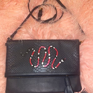 Pode incluir: Clutch preta de pele de cobra com um design de cobra vermelho e branco. A bolsa tem um fecho de correr e um tassel.
