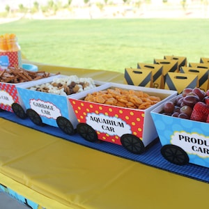 Puede incluir: Una mesa de comida para una fiesta con temática de tren, con cinco cajas decoradas etiquetadas como "Produce Car", "Aquarium Car", "Garbage Car", "Log Car" y "Grandma Car". Las cajas están llenas de bocadillos como pretzels, galletas saladas y fruta.