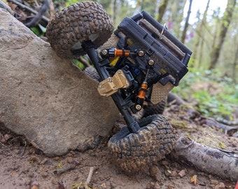 May include: A black and gold toy truck with orange suspension and black tires is on a rock in a forest setting.
