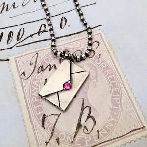 Peut inclure: Un collier en argent avec un petit pendentif en forme d'enveloppe en argent orné d'une pierre précieuse rose. Le collier repose sur un timbre-poste vintage avec le texte "Jan" et "One Penny".